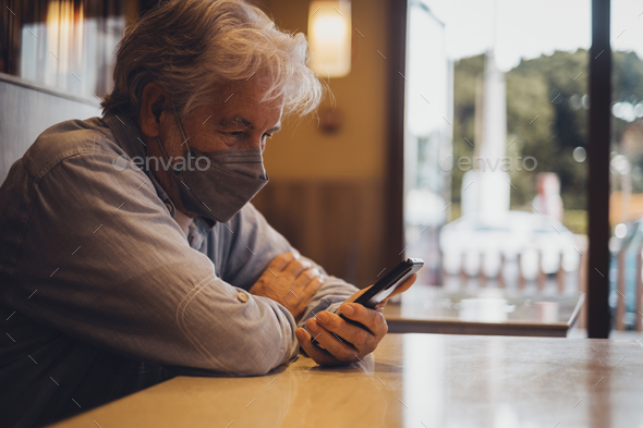 Senior man sitting at the table looking worried mobile phone and wearing protective mask