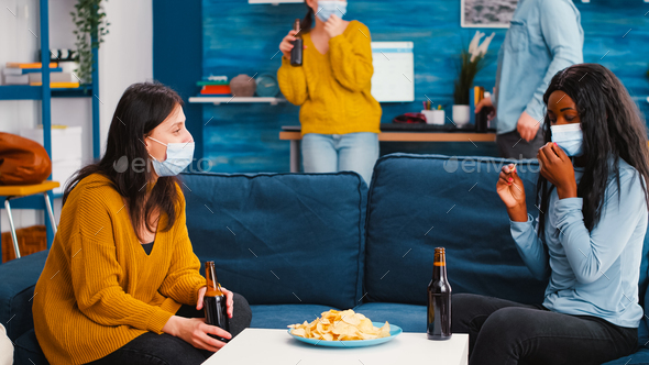 Women sitting on sofa discussing taking off protective mask eating snacks
