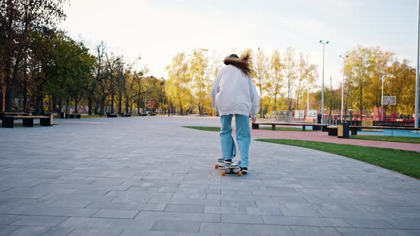 Rear View of a Young Stylish Female Student Riding a Longboard in the Park alt