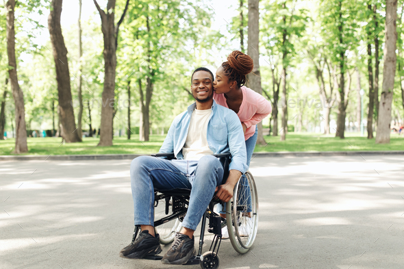 Young loving black woman kissing her happy boyfriend in wheelchair on walk at city park, full length
