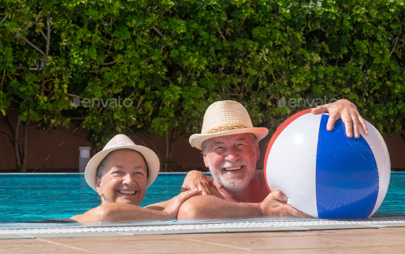 Happy senior couple laughing floating in outdoor swimming pool playing with inflatable balloon