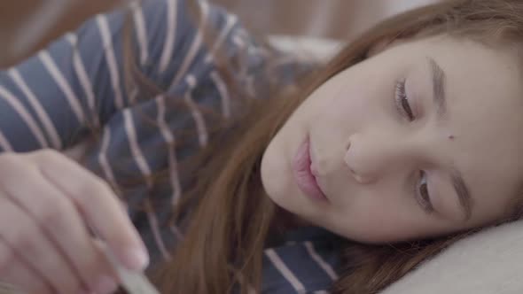 Teenage Girl Wrapped in a Blanket Lying in Bed Checking Temperature on Thermometer Close Up alt