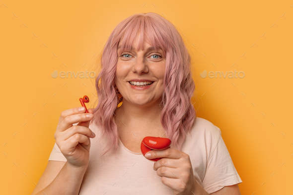 caucasian woman with pink curly hair inserting wireless earphone in ear posing