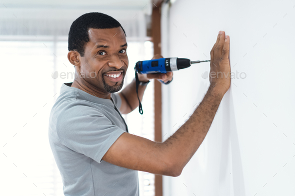 African American man builder drilling hole in a new white wall in new house with drill