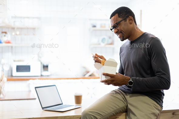 African American man eating noodle in a take away box and coffee cup at home while sit using laptop