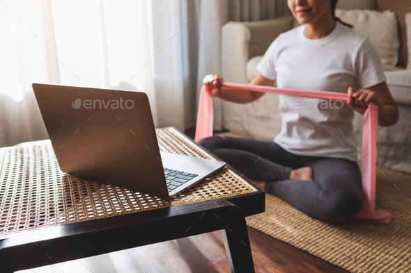 A woman holding rubber resistance band while watching online workout tutorials on laptop at home