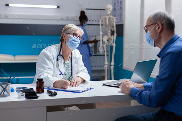 Woman doctor with face mask taking notes on papers at medical consultation with man