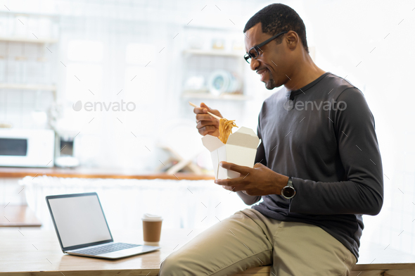 African American man eating noodle in a take away box and coffee at home while sitting using laptop
