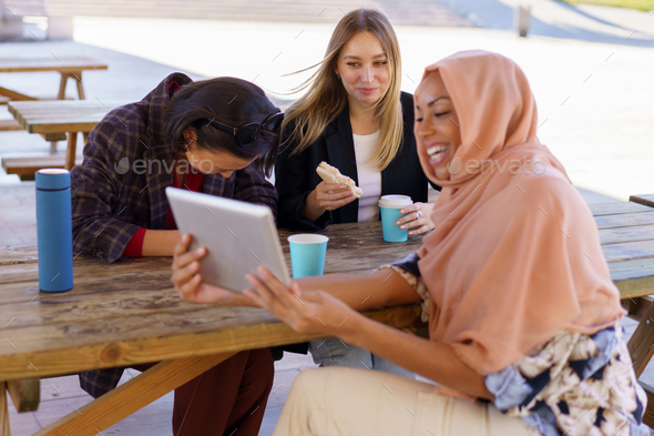 Content young multiracial female friends laughing while sharing tablet in cafe