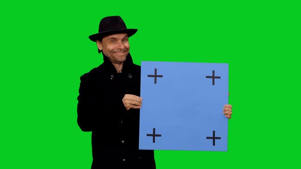 Smiling Man In Black Hat And Coat Looking At Camera And Pointing Blue Mockup Board
