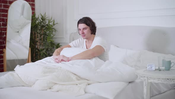 Young Brunette Man Yawning and Stretching Sitting on Bed in the Morning alt