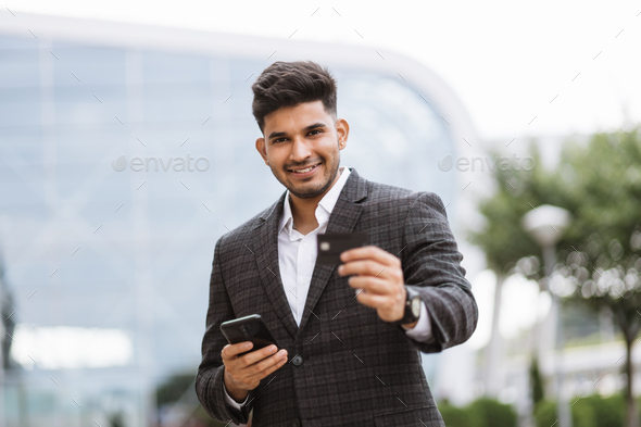 Businessman stands outside at airport terminal, uses mobile cell phone and credit bank card