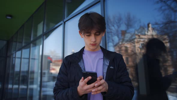 Medium Shot of Handsome Young Man Working on Smartphone Stands on the Street in Front of a Modern
