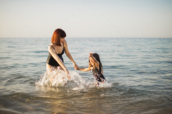 Young red hair mother in black bikini standing in water and playing with her small smiling daughter