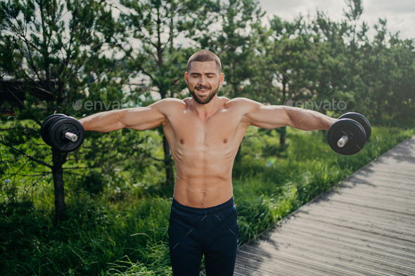 Fit adult man lifts heavy weight during workout session outdoor, poses with naked torso