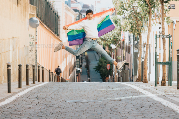 Young man holding a gay pride flag jumping at the street. Styling and liberty concepts