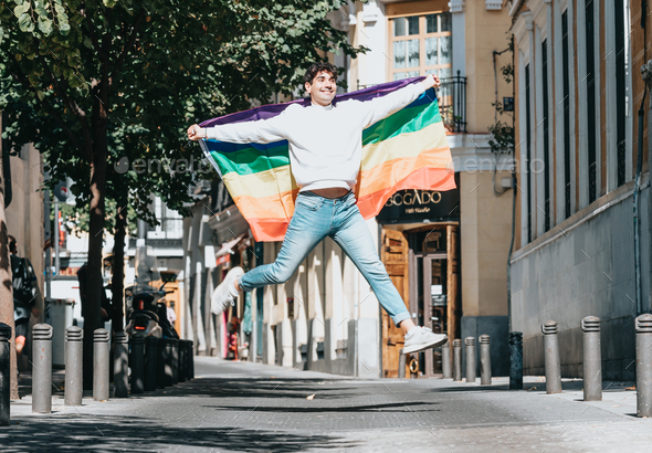 Young man holding a gay pride flag jumping at the street. Styling and liberty concepts.