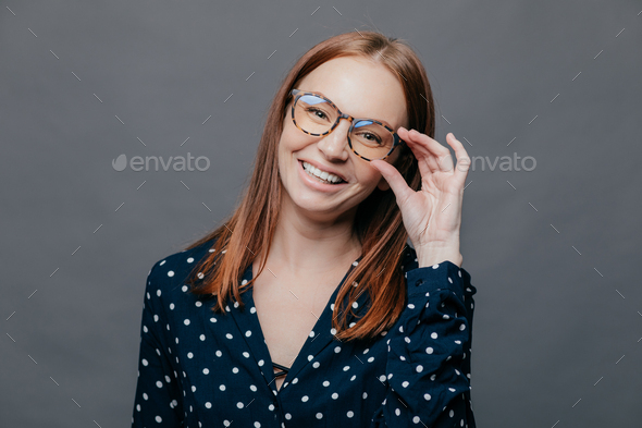 Smiling woman with pleased facial expression, keeps hand on rim of spectacles, wears black polka dot