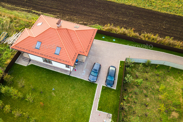 Aerial top view of house shingle roof with attic windows and cars on paved yard with green grass