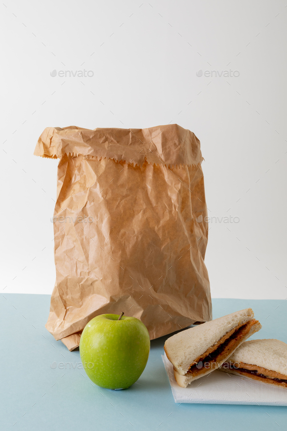 Green apple with paper bag and peanut butter and jelly sandwich on table against gray background