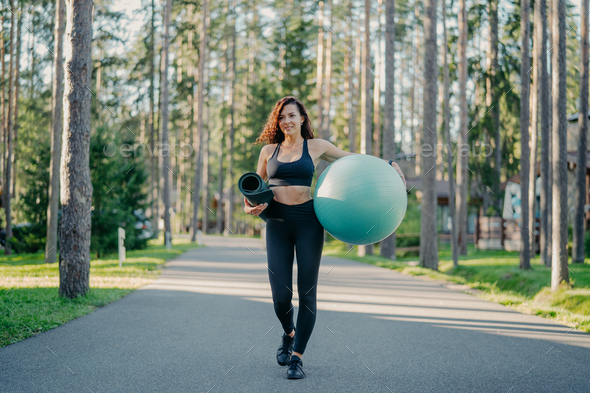 Active slim woman dressed in sportswear, carries fit ball and rolled up karemat, walks on road