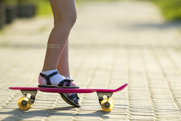 Child slim legs in white socks and black sandals on plastic pink skateboard on bright sunny summer