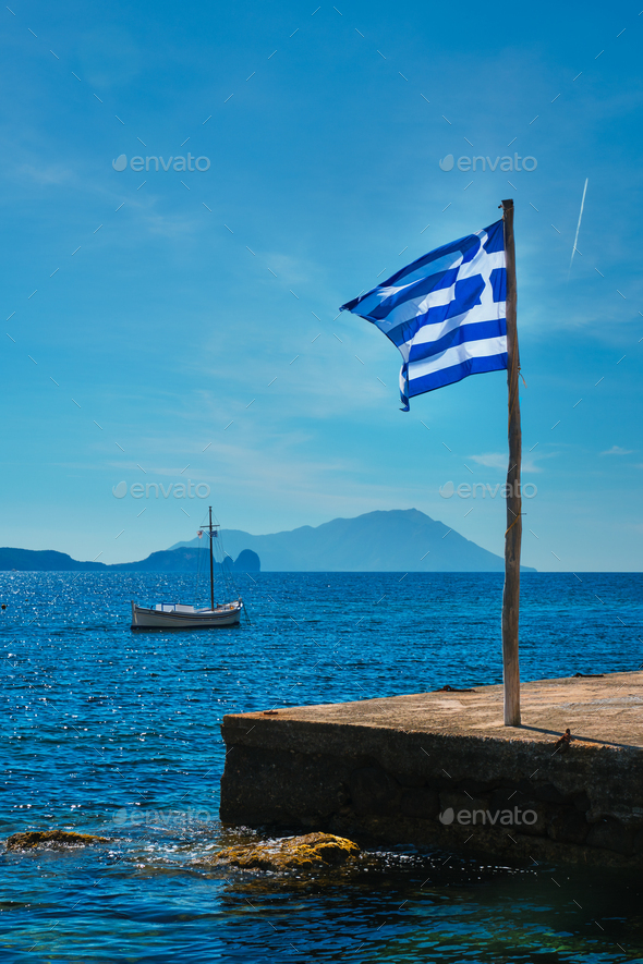 Greek flag in the blue sky on pier and traditional greek fishing boat in  the Aegean sea Stock Photo by Dmitry_Rukhlenko