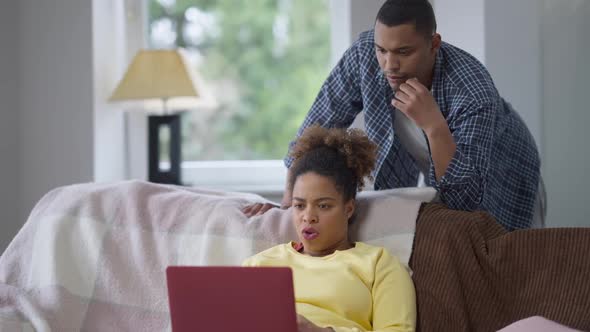 Focused African American Woman Messaging Online on Laptop As Worried Man Supporting Wife Talking and alt