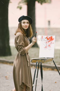 Girl in dress and black hat painting on an easel in a park