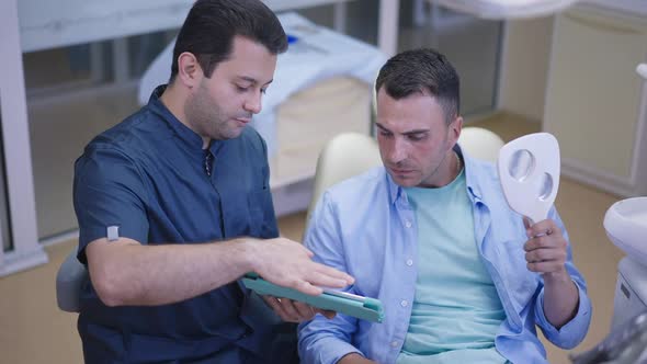 Handsome Caucasian Man Examining Dentist Work in Mirror As Middle Eastern Doctor Talking in Slow alt