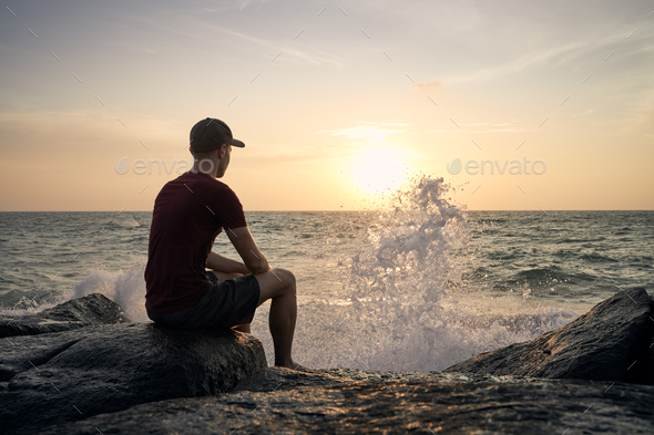 Man sitting on rock and watching sunset over ocean Stock Photo by Chalabala