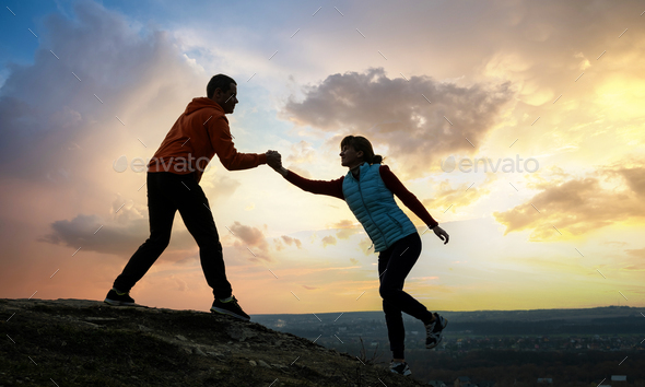 Man and woman hikers helping each other to climb stone at sunset in mountains. Couple climbing on