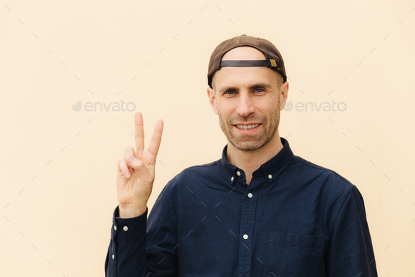 Handsome delighted unshaven male shows peace sign, keeps two fingers raised