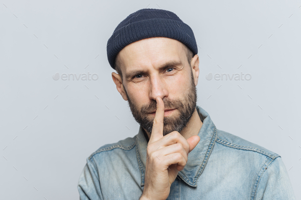 Handsome man with thick beard and mustache, has secret look, demonstrates silence sign