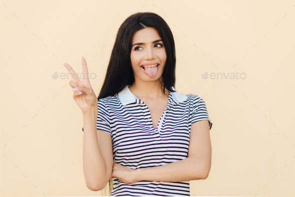 Positive funny female makes peace sign with hand, shows tongue, being in good mood