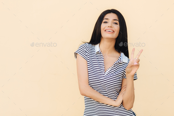 Cheerful brunette female shows peace gesture, keeps two fingers raised, has pleasant smile