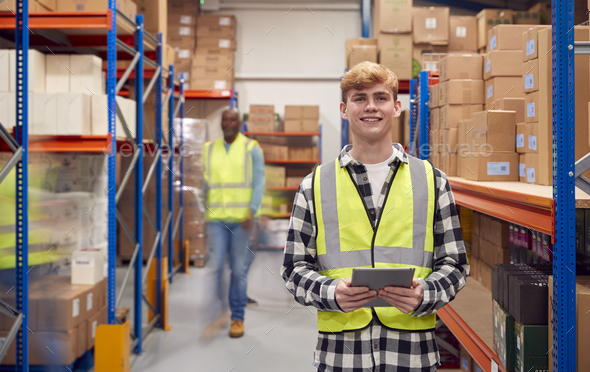 Portrait Of Male Intern With Digital Tablet In Busy Modern Warehouse With Staff In Background