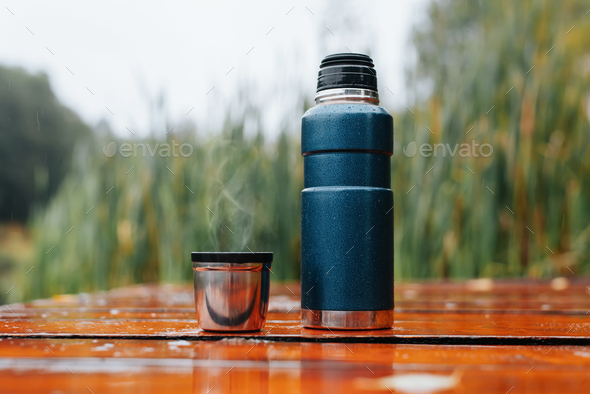 Hike, camping concept. Thermos and aluminum mug hot drink with rising steam  standing on stump in Stock Photo by SergioPhotone