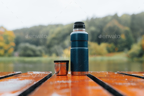 Thermos and aluminum mug with hot drink and rising steam outdoors. Camping vacuum flask bottle