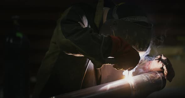 Welder in welding mask, in working clothes at the factory, welds the pipe in the factory room.  alt