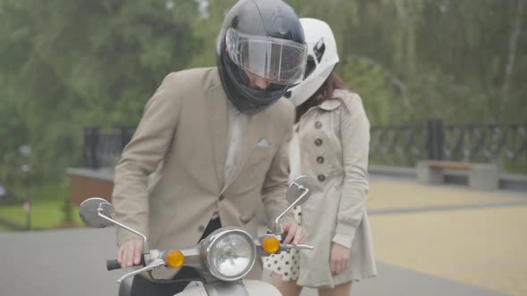 Caucasian Couple of Bikers in Helmets Coming To Scooter Outdoors and Sitting Down on It. Confident alt