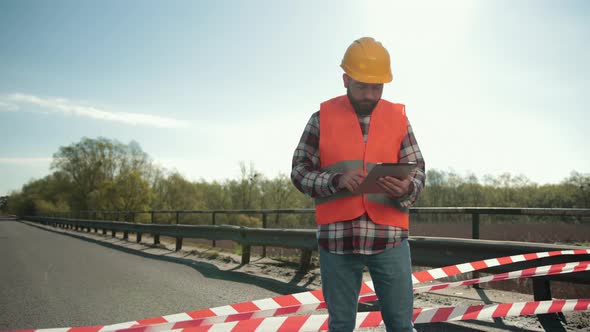Young Bearded Man Inspector in an Orange Vest and Protective Helmet with Tablet alt