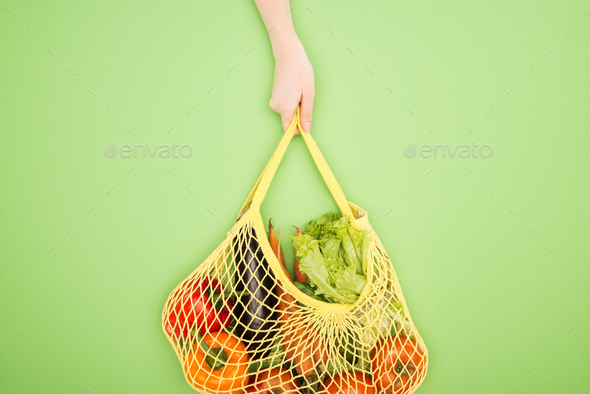 partial view of woman holding string bag with whole vegetables on light green