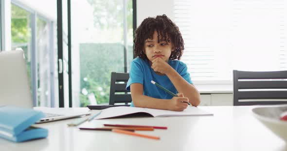 Happy biracial boy sitting at table in kitchen doing homework alt
