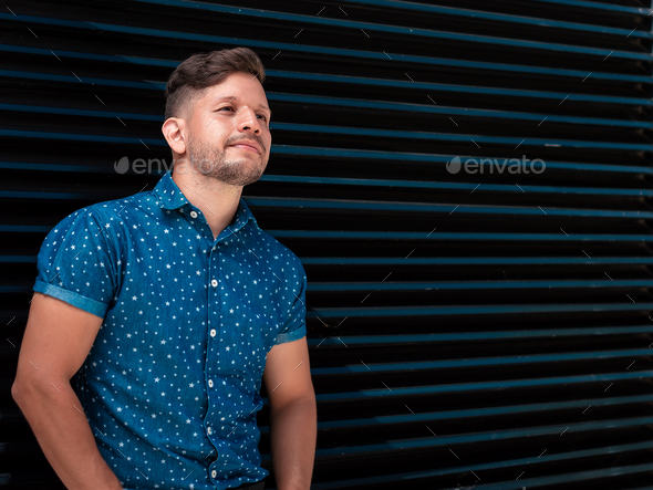 young latin guy, with beard and short hair, leaning against a blue and black metal wall.