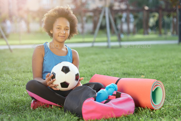 Cheerful little girl with soccer ball sitting on grass outdoors
