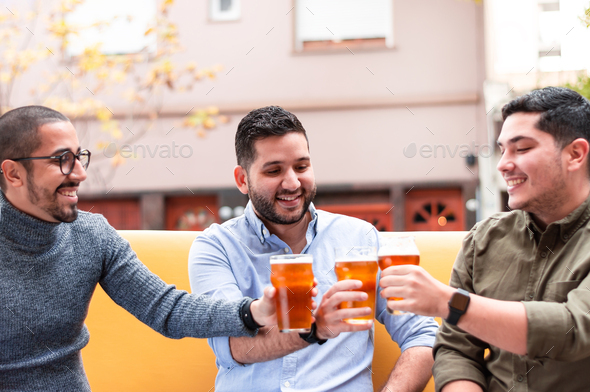 Group of Latino friends, toasting and smiling, drinking a craft beer outside a bar.