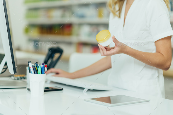 Pharmacist holding medical cream and using computer while working at a pharmacy