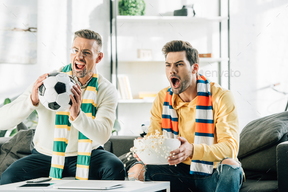 excited son and father with scarfs watching football and screaming at home