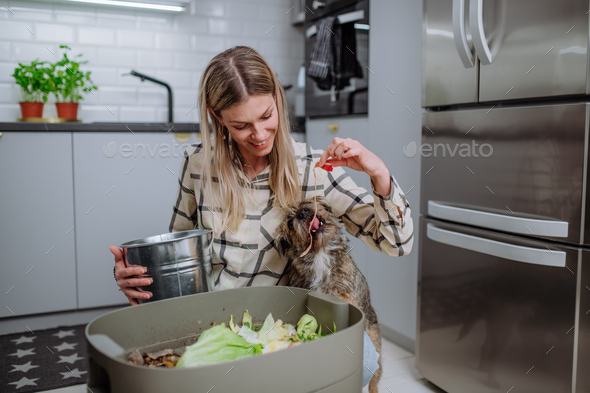 Woman throwing vegetable cuttings in a compost bucket in kitchen and feeding dog